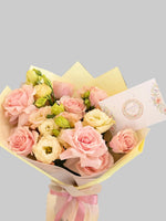 Load image into Gallery viewer, A bouquet of pink and cream roses wrapped in pale yellow paper with a pink ribbon, accompanied by a floral card, set against a blurred indoor background.